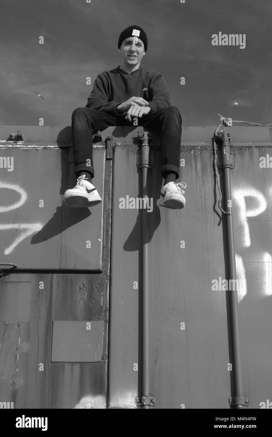 Black and White Industrial Portrait of a Young Man Atop an Old Shipping Container at Cruden Bay Harbour, Aberdeenshire, Scotland, UK. Winter, 2018. Stock Photo