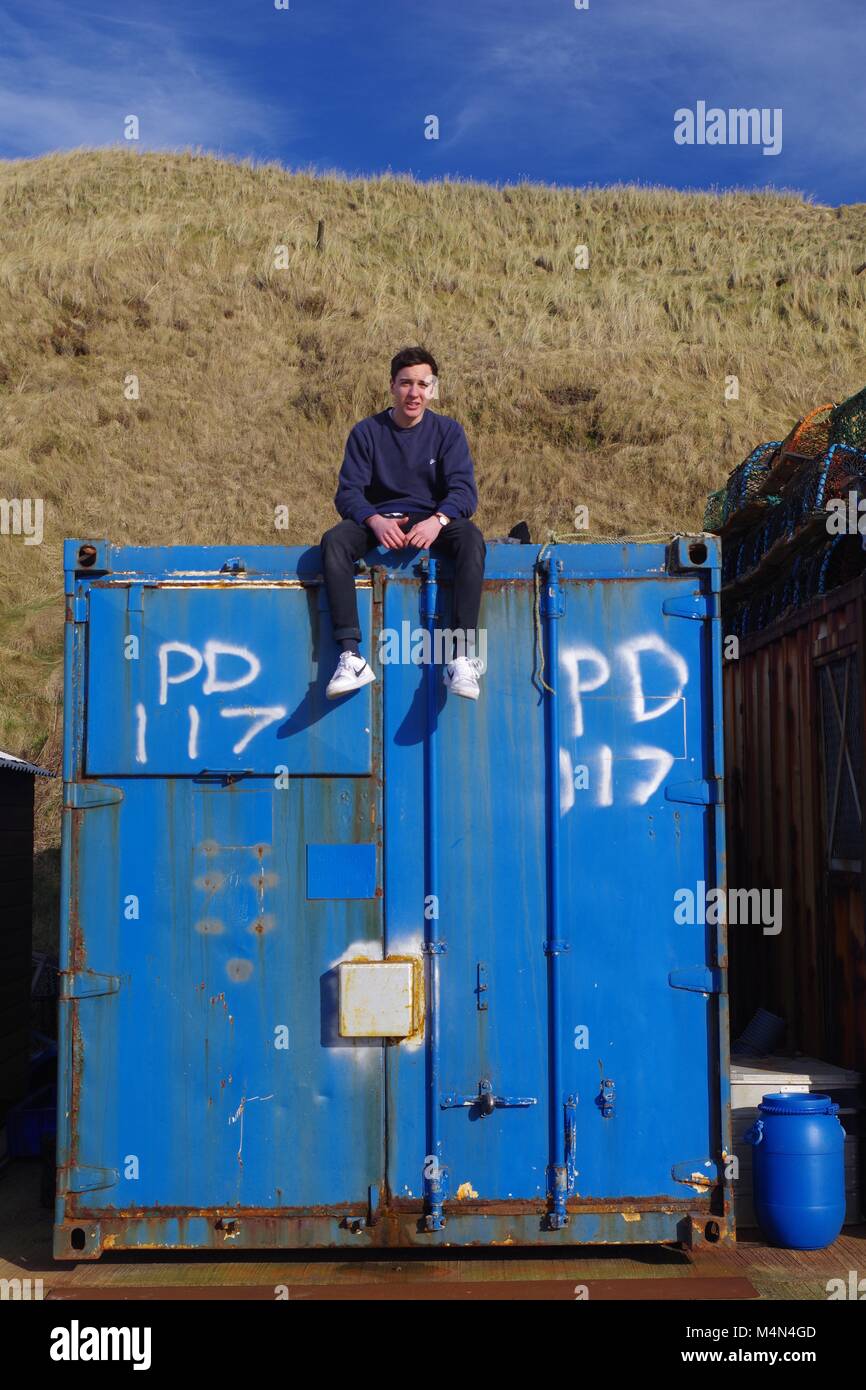 Young Man Sitting Atop an Old Blue Shipping Container at Cruden Bay