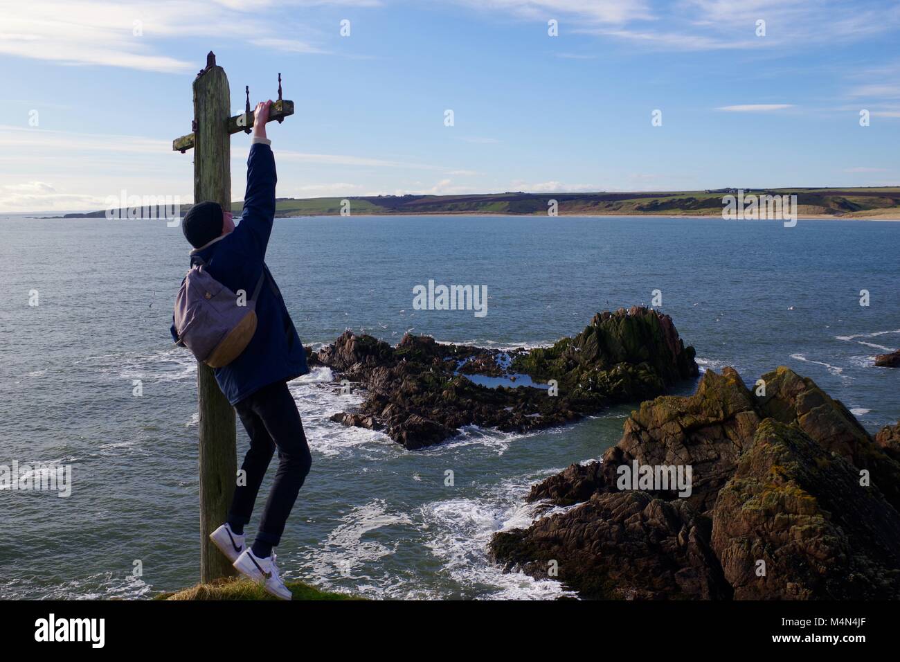 Young Adventurous Man Dangles from an Old Telegraph Pole at the Edge of a Liff. Cruden Bay, Aberdeenshire, Scotland, UK. Feb, 2018. Stock Photo
