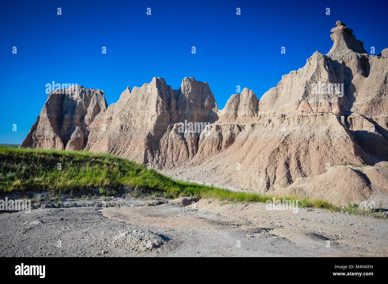 Horizontal view of painted desert in Badlands National Park shows layers of bright blue sunny sky, red and white striped hills, green grass, and grave Stock Photo