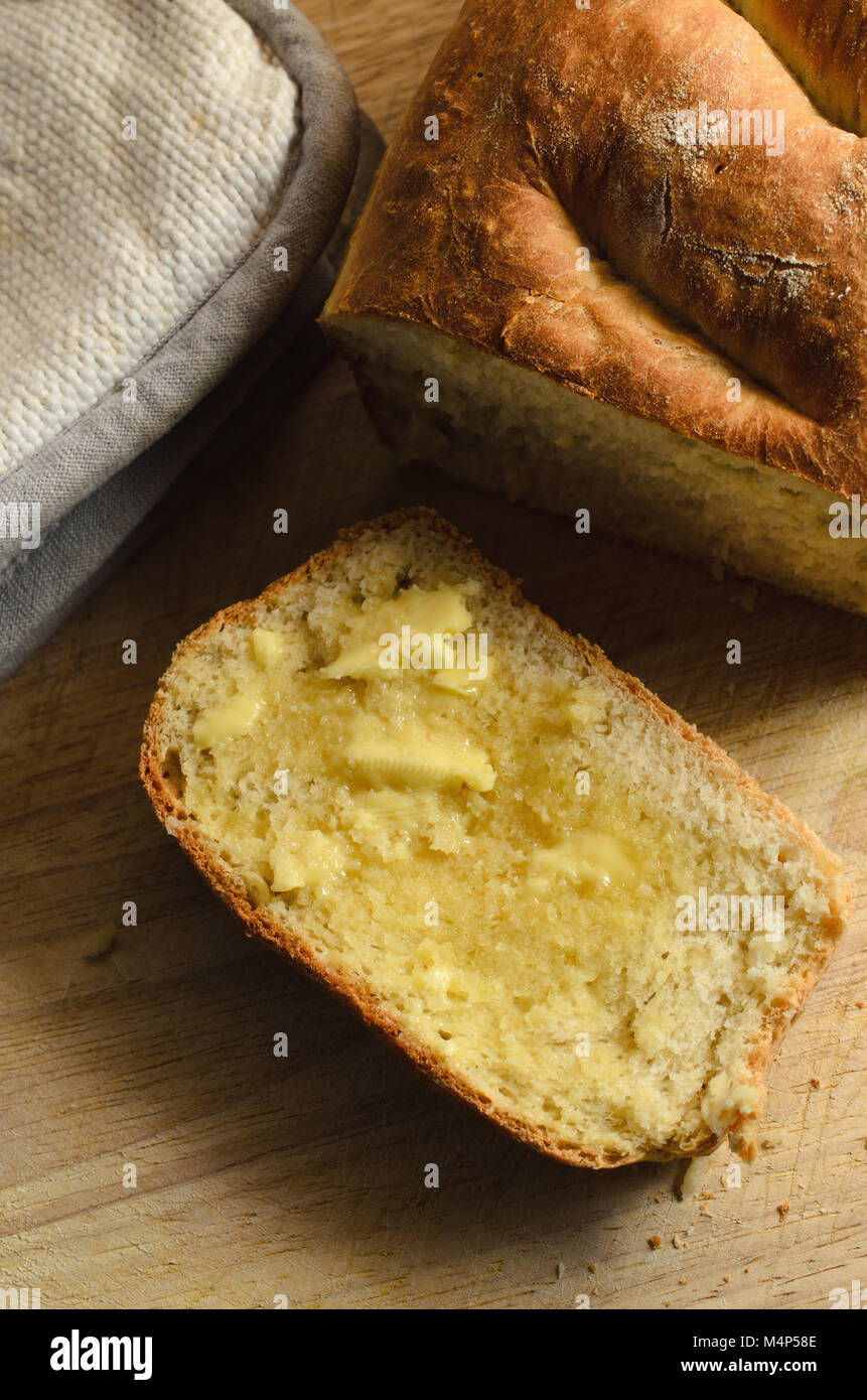 Overhead shot of a crusty, home baked loaf of bread with butter melting into still warm upturned slice. On wooden board with crumbs. Stock Photo