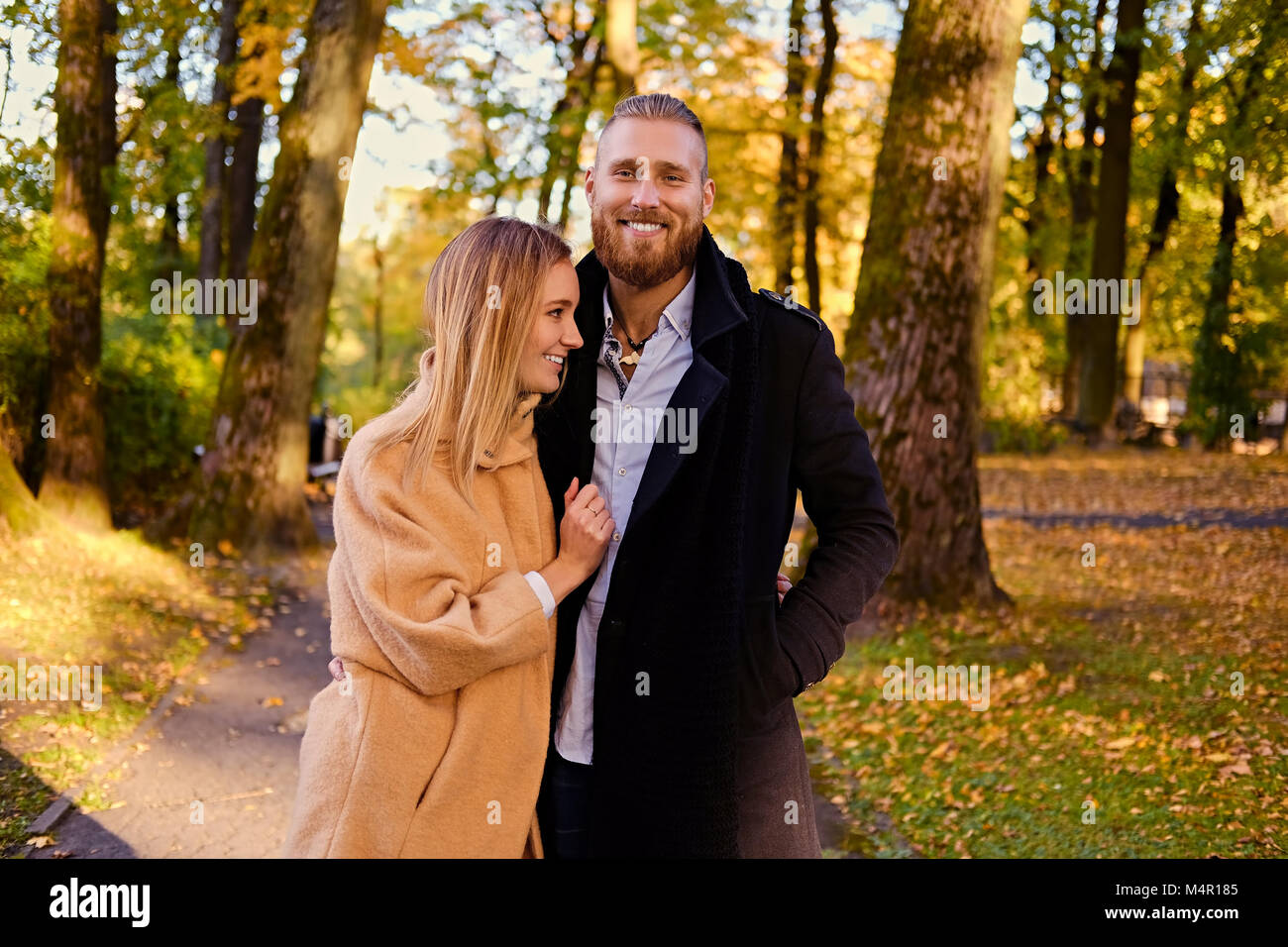 Autumn Love story. Redhead bearded male hugs the cute blonde female on the date in an autumn park. Stock Photo