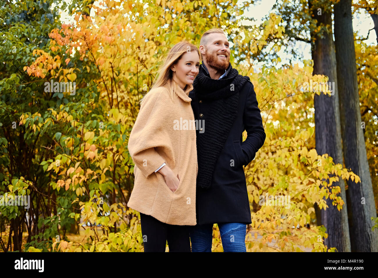 Autumn Love story. Redhead bearded male hugs the cute blonde female on the date in an autumn park. Stock Photo