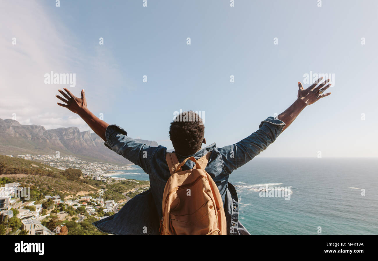 Rear view of young guy with backpack standing outdoors with arms spread open against seascape. Man enjoying the view from the top of the mountain. Stock Photo