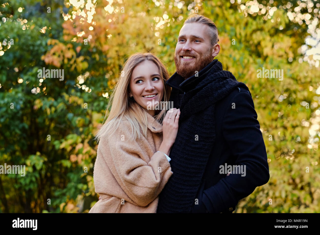 Autumn Love story. Redhead bearded male hugs the cute blonde female on the date in an autumn park. Stock Photo