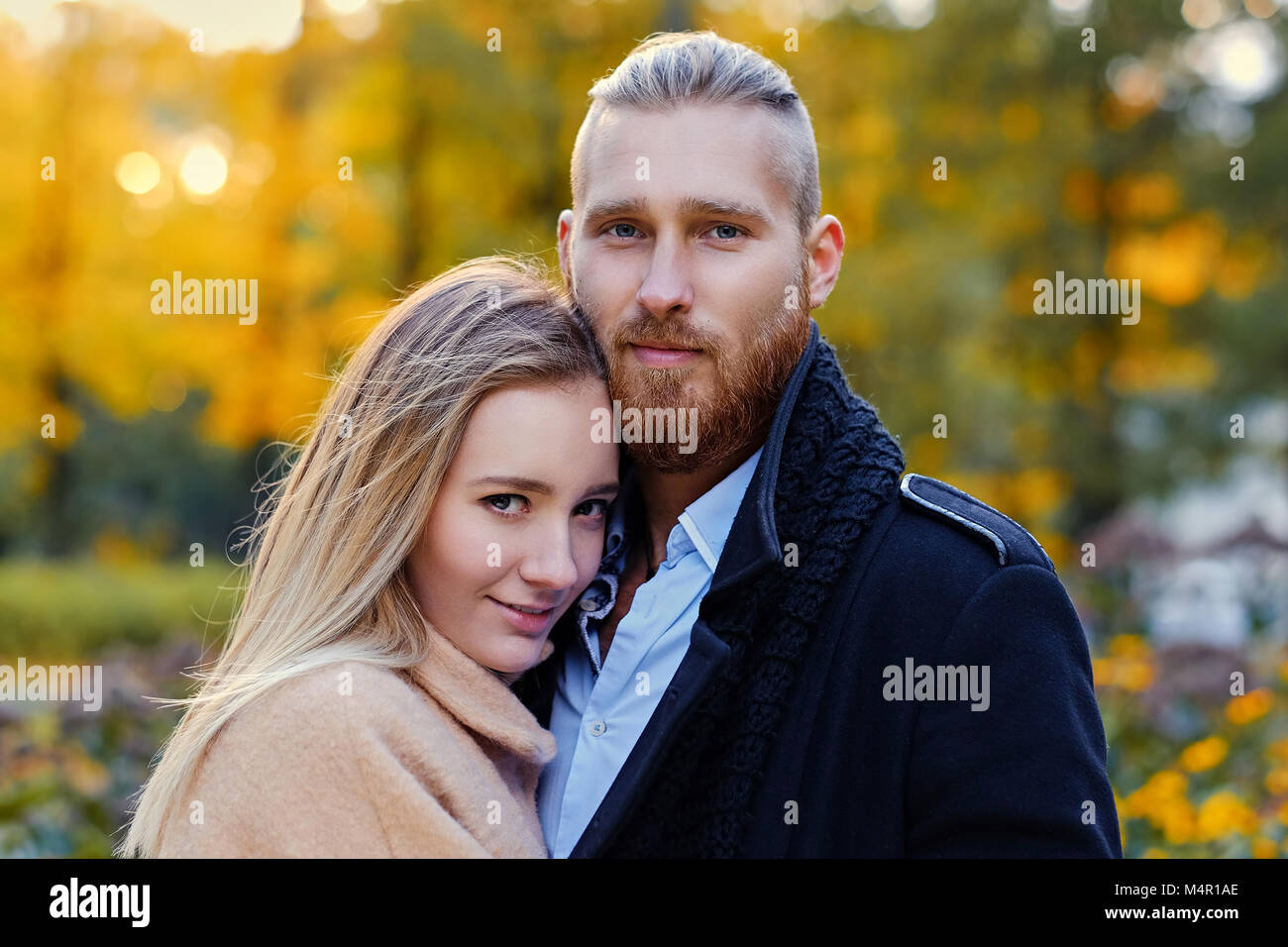 Close up portrait of a couple on a date walk in autumn park. Positive redhead bearded male hugs the cute blonde female. Stock Photo