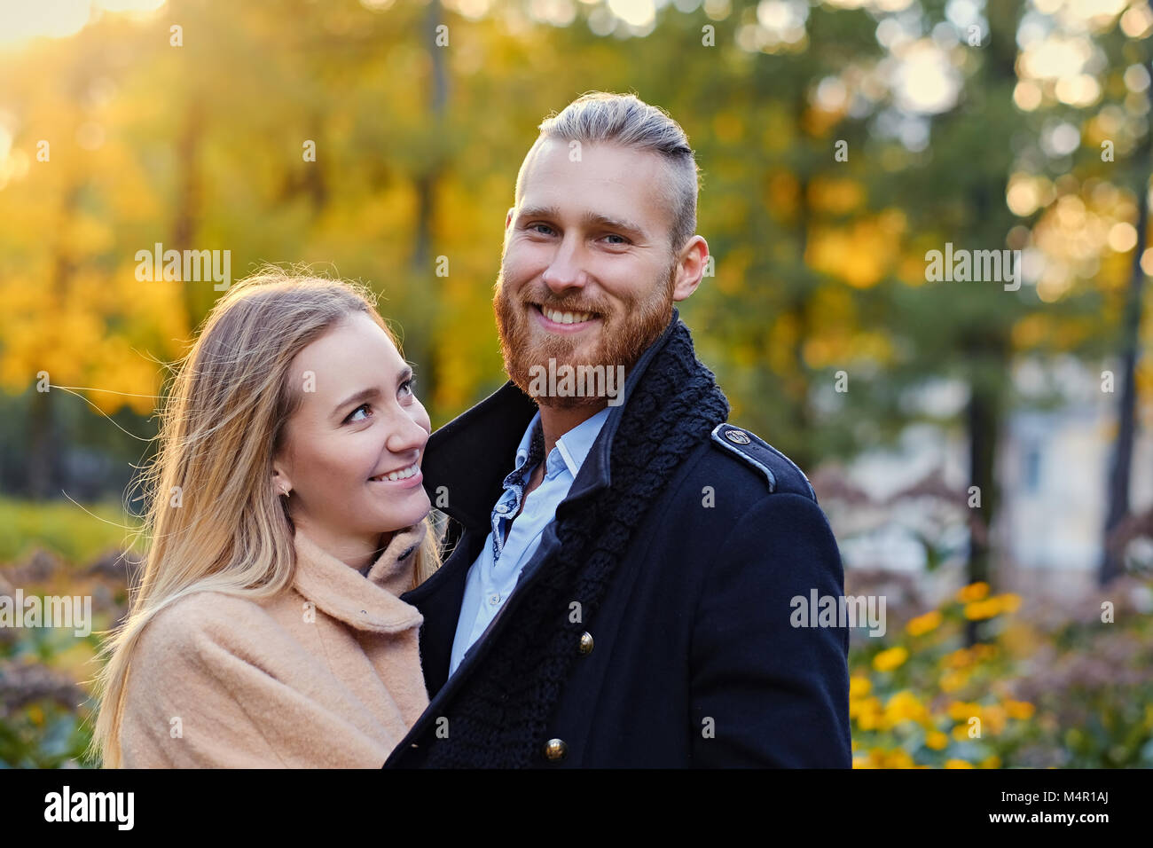 Close up portrait of a couple on a date walk in autumn park. Positive redhead bearded male hugs the cute blonde female. Stock Photo