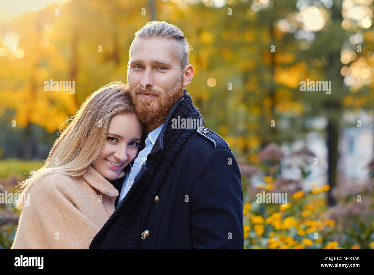 Close up portrait of a couple on a date walk in autumn park. Positive redhead bearded male hugs the cute blonde female. Stock Photo