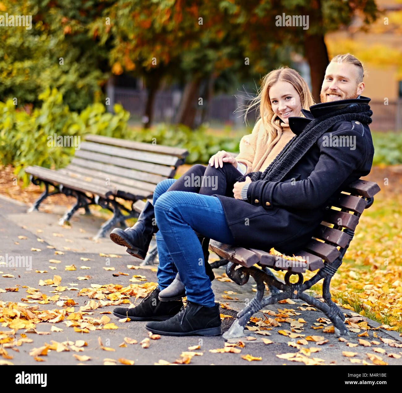 Positive smiling couple of redhead male and blonde female relaxing on a bench in an autumn city park. Stock Photo