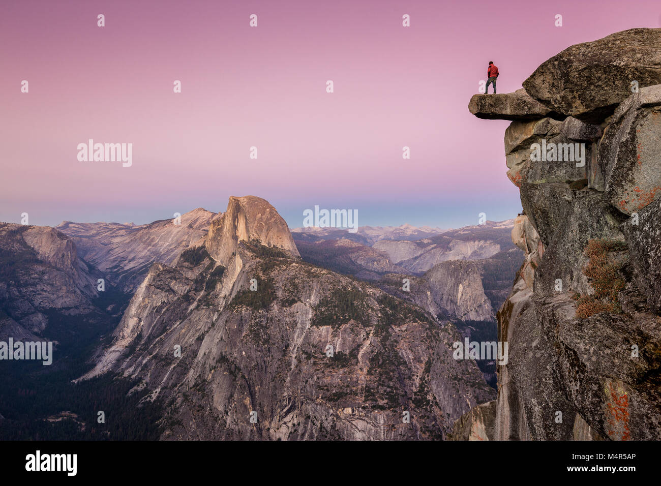 A fearless male hiker is standing on an overhanging rock at Glacier Point enjoying the breathtaking view towards famous Half Dome in beautiful post su Stock Photo