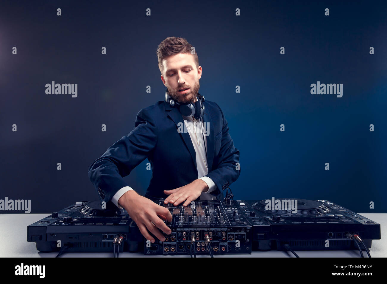 Closeup portrait of confident DJ with stylish hair style and headphones on neck mixing music on mixer while standing isolated on dark colored blue, cyan background Stock Photo