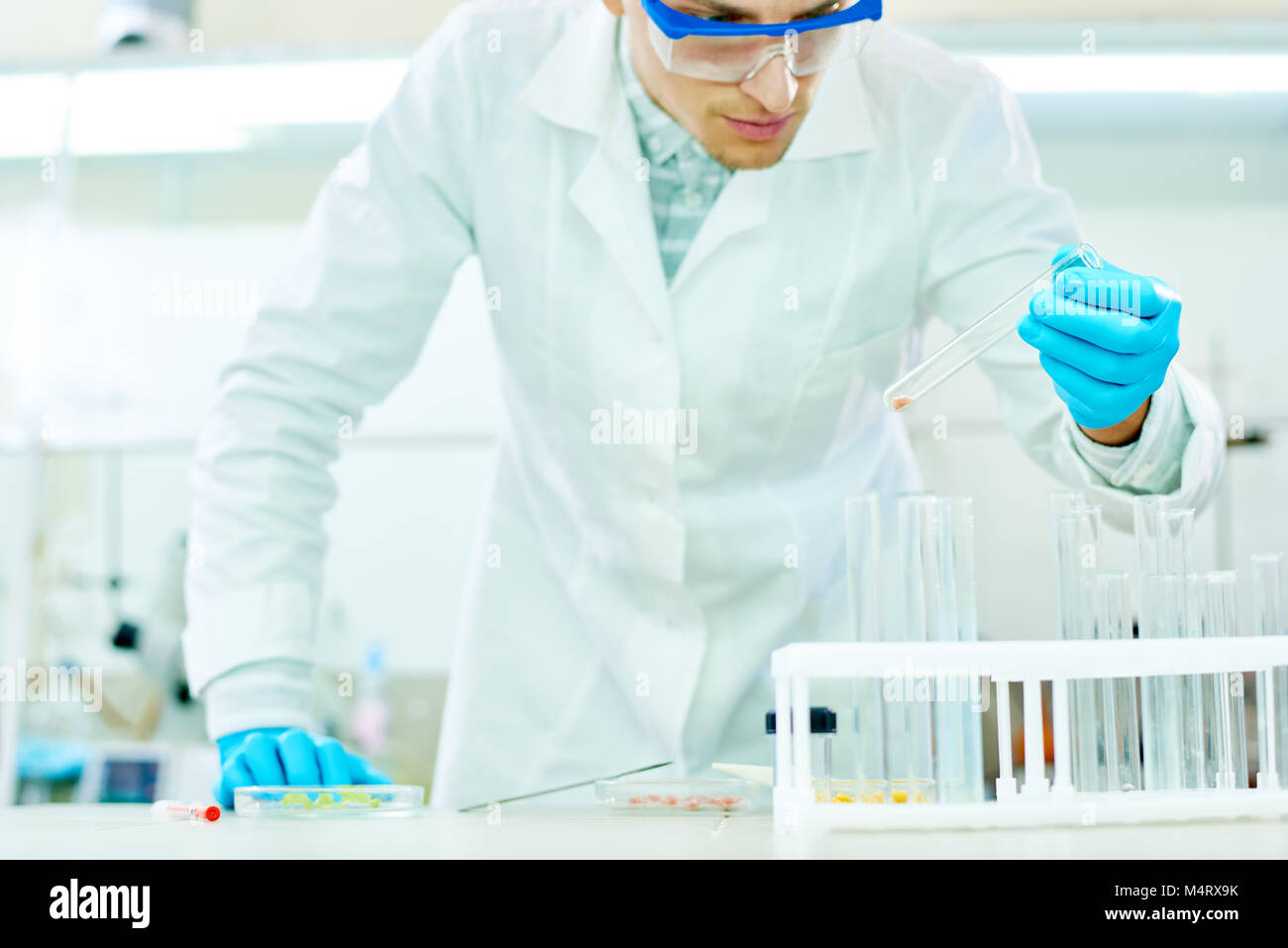 Handsome young researcher wearing safety goggles and white coat looking at test tube with concentration while carrying out experiment in modern lab Stock Photo