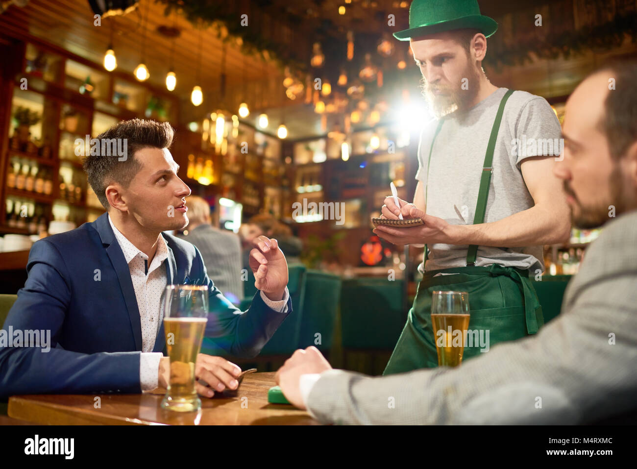 Celebrating St. Patricks Day in cafe: two friends wearing suits sitting at table and making order while bearded waiter in leprechaun costume standing  Stock Photo