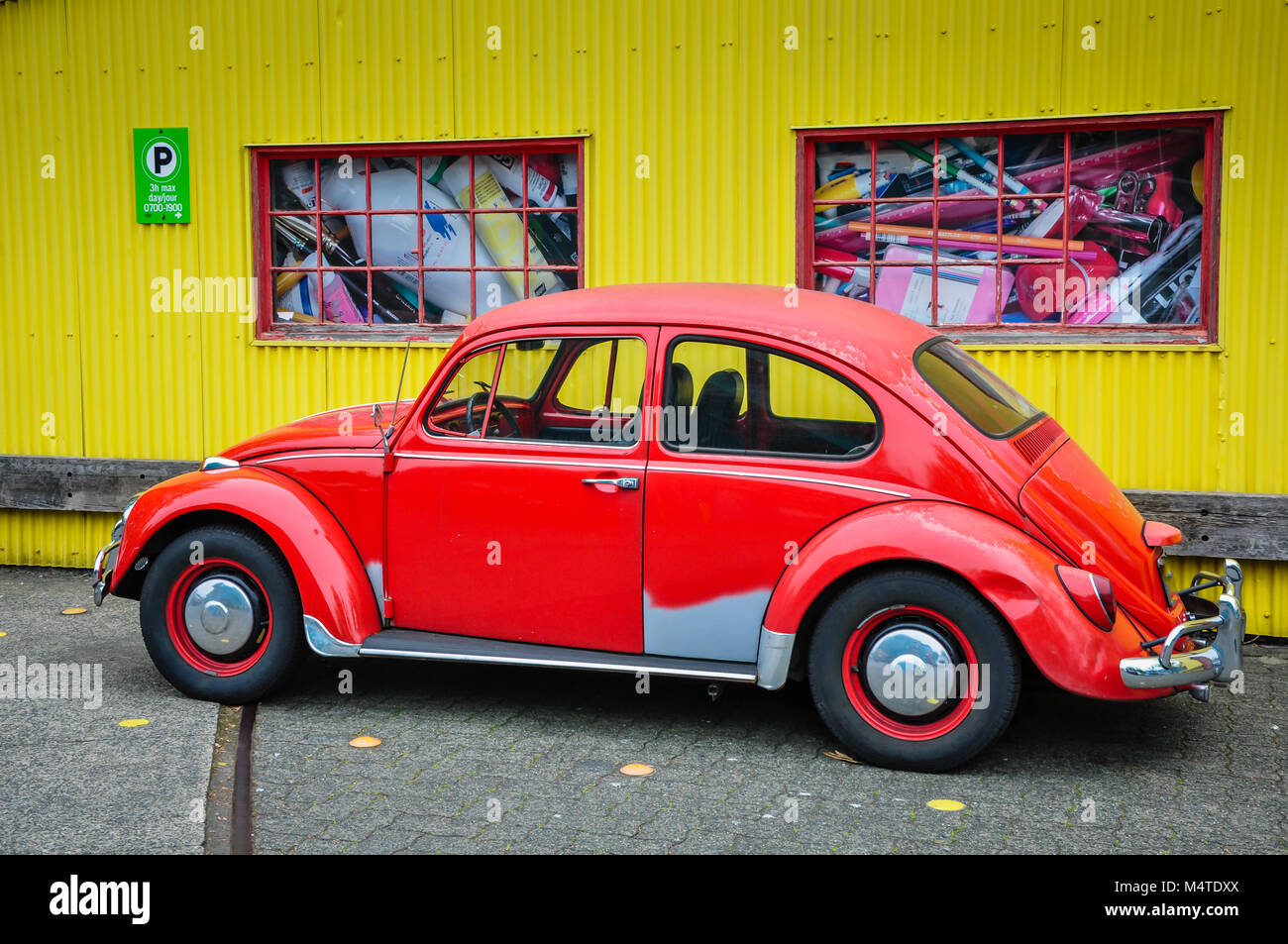 Vintage Volkswagon Bug car mostly painted red, parked in front of yellow corrugated tin wall. Stock Photo