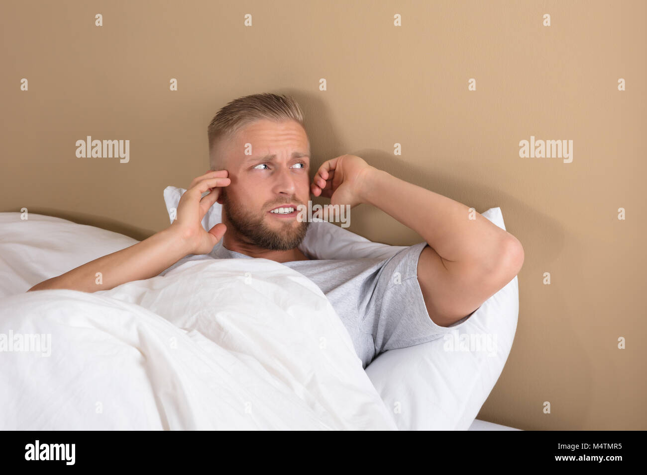 Troubled Young Man Lying On Bed Covering His Ears To Avoid Noise Stock Photo