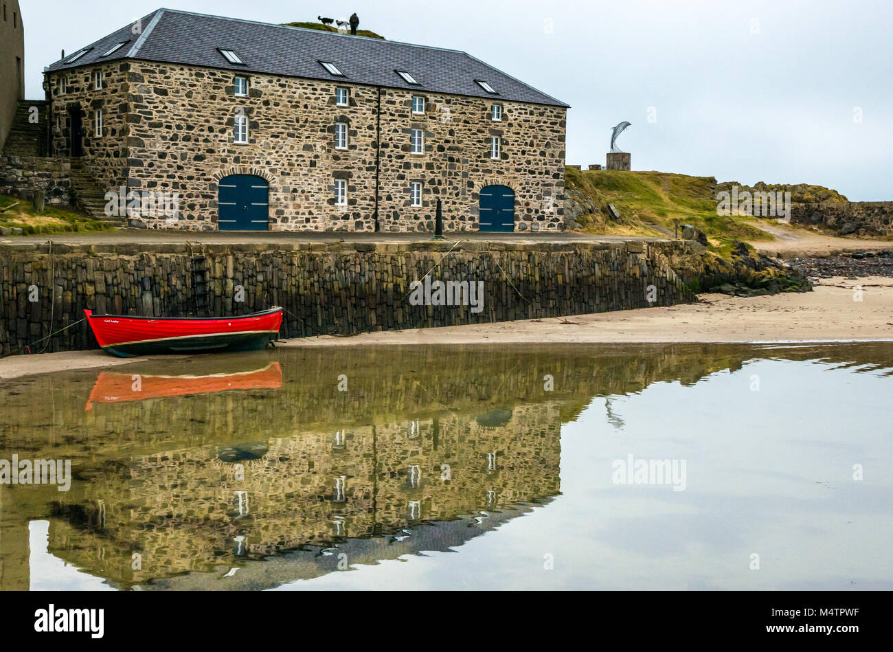 Small red rowing boat, picturesque harbour, Portsoy, Aberdeenshire, Scotland, UK, with water reflection, historic buildings dolphin sculpture Stock Photo