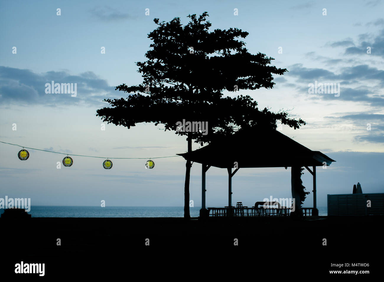 Silhouettes of a big tree and gazebo on the coast in the evening and a garland with round yellow lights. Bali, Indonesia. Stock Photo