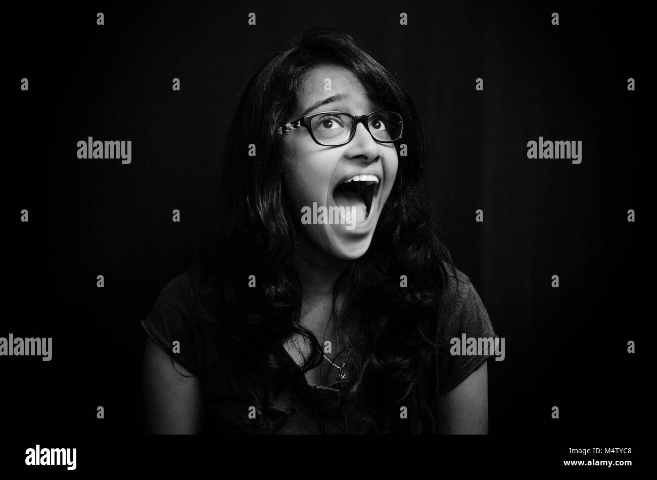 Black and white portrait of a young adult girl with eyeglasses, screaming out of joy, uncontrolled burst of emotion over black background. Stock Photo