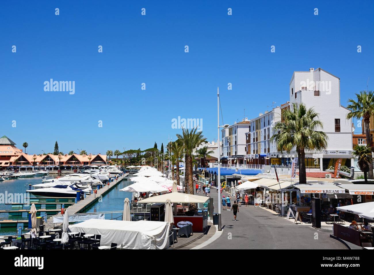 Boats moored in the marina with tourists walking along the restaurant lined promenade, Vilamoura, Algarve, Portugal, Europe. Stock Photo