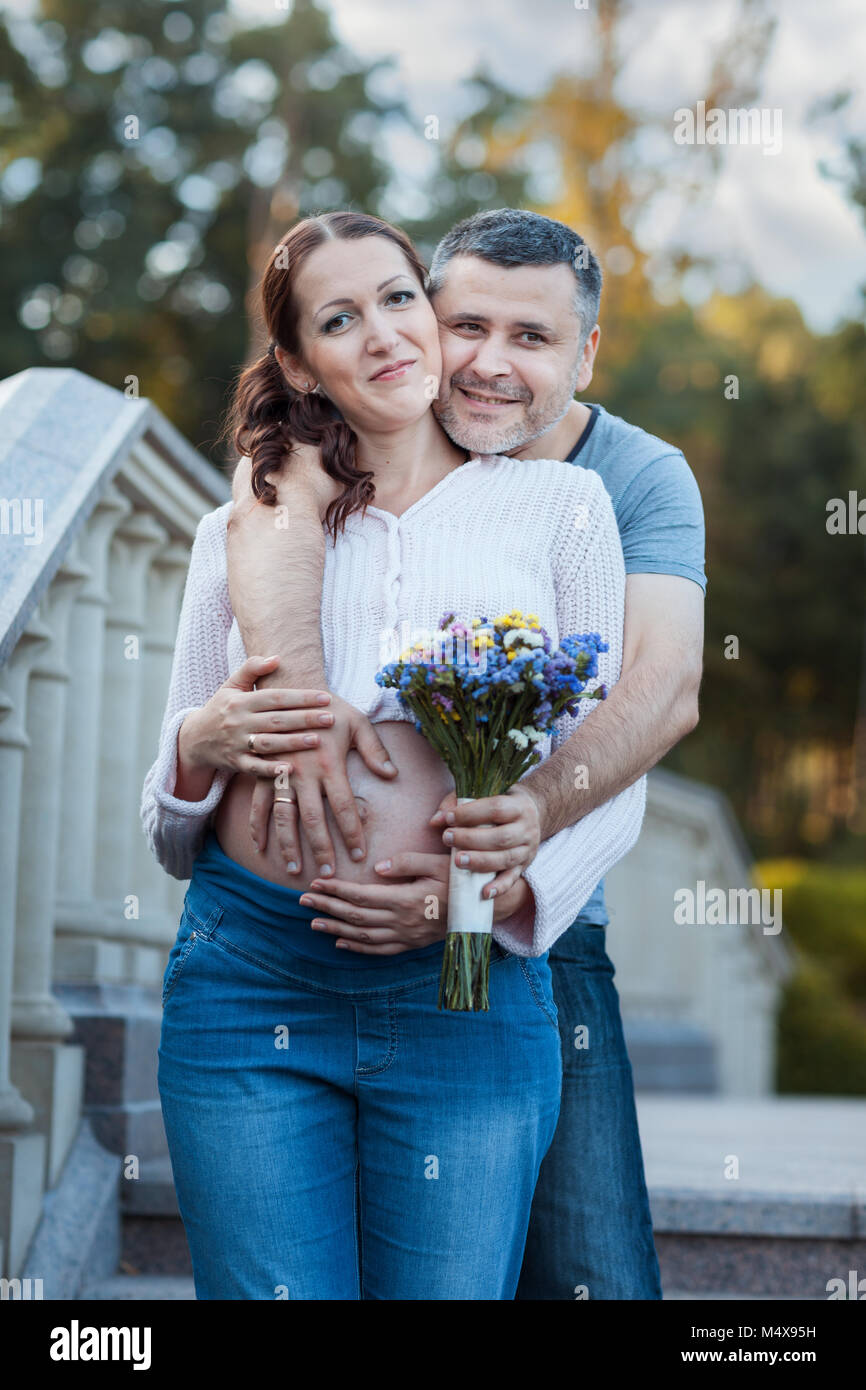 Young man embracing his pregnant wife. He holds a bouquet of flowers in ...
