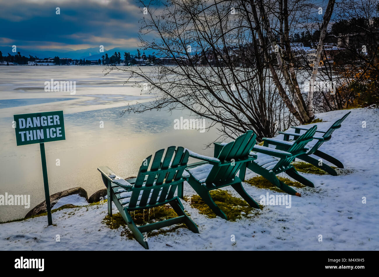 Posted sign warning of thin ice on Mirror Lake in Lake Placid, NY. Four green Adirondack chairs line the edge of the lake, next to white birch trees.  Stock Photo