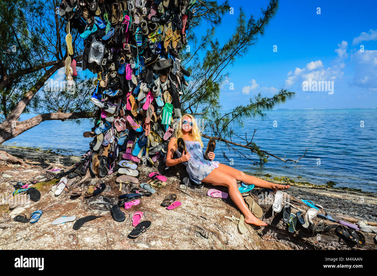 Sun-tanned blonde girl rests against a tree covered in flip flops left by tourists in Grand Cayman island in the Caribbean. Stock Photo