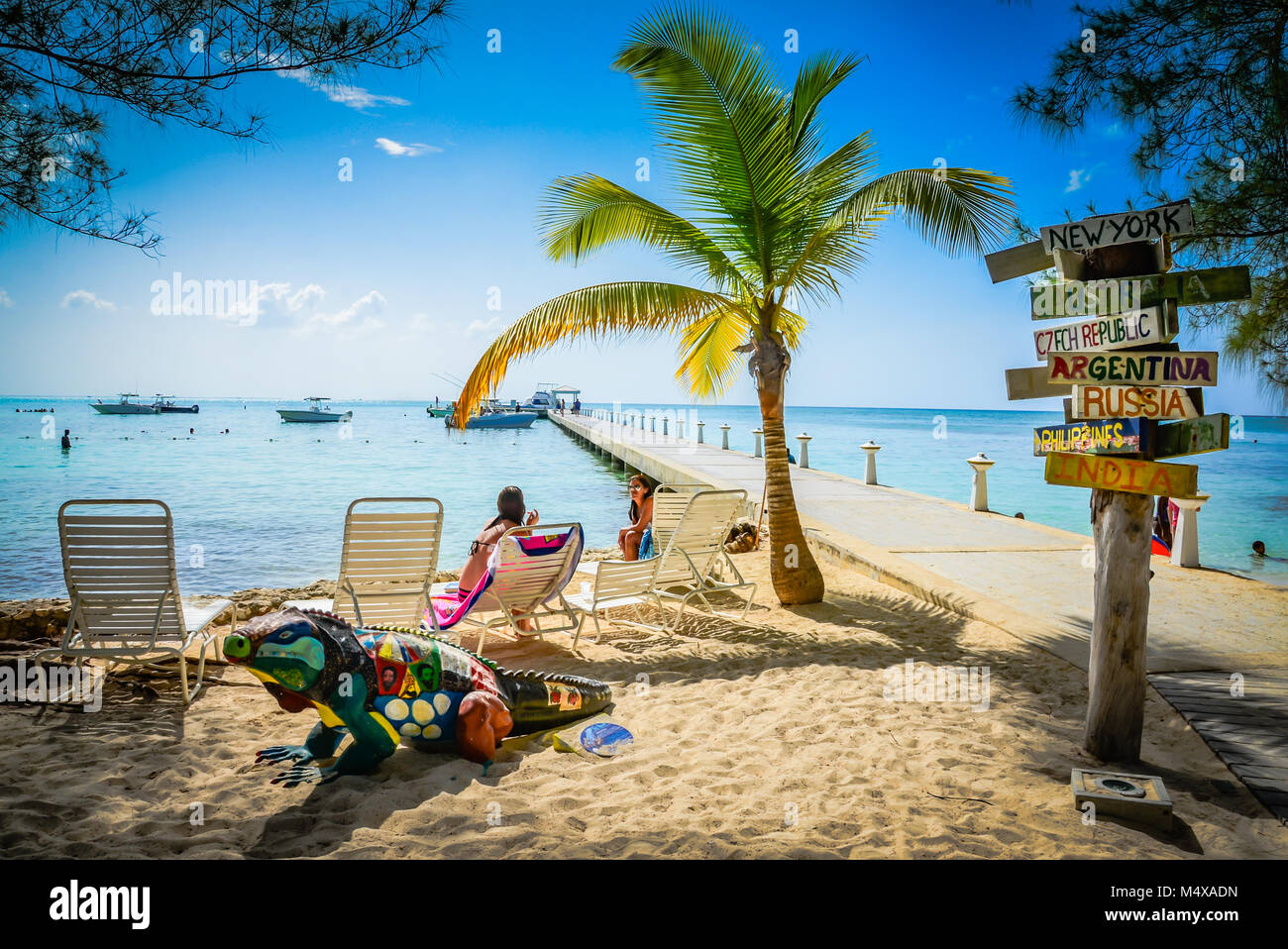 International destinations signpost at pier next to chaise lounges and palm trees on beach with blue iguana statue at Rum Point Club, Grand Cayman. Stock Photo