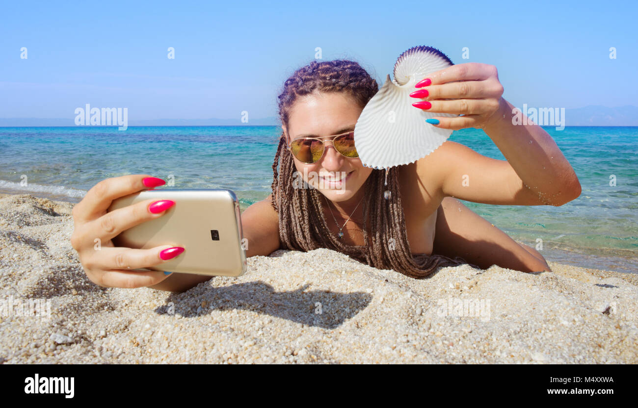 Gorgeous female is making selfie with smartphone lying on a sandy beach of a beautiful distant sea shore, holding nautilus shell. Stock Photo