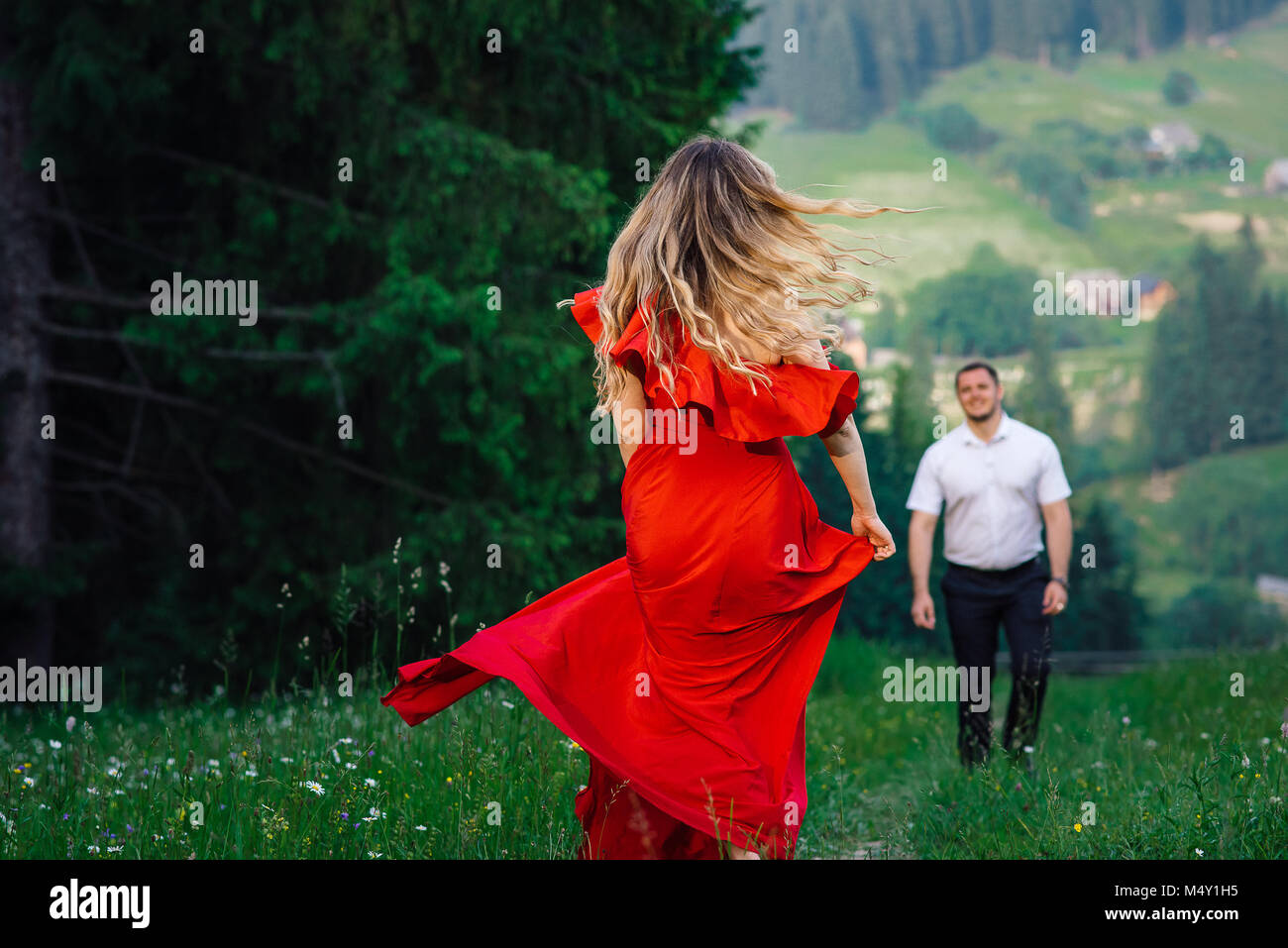 Back view of the elegant woman in long red dress with blonde curly hair spinning round at the blurred background of her groom in the green mountains. Stock Photo