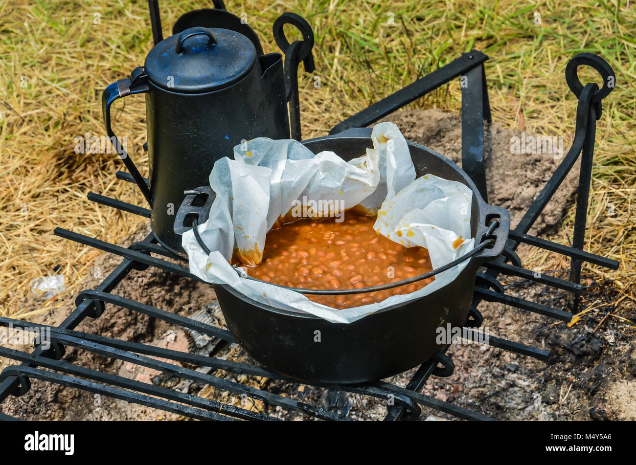 Traditional Boston Baked Beans cooked over hot coal in a black cast iron pot, with a kettle on the side. Stock Photo
