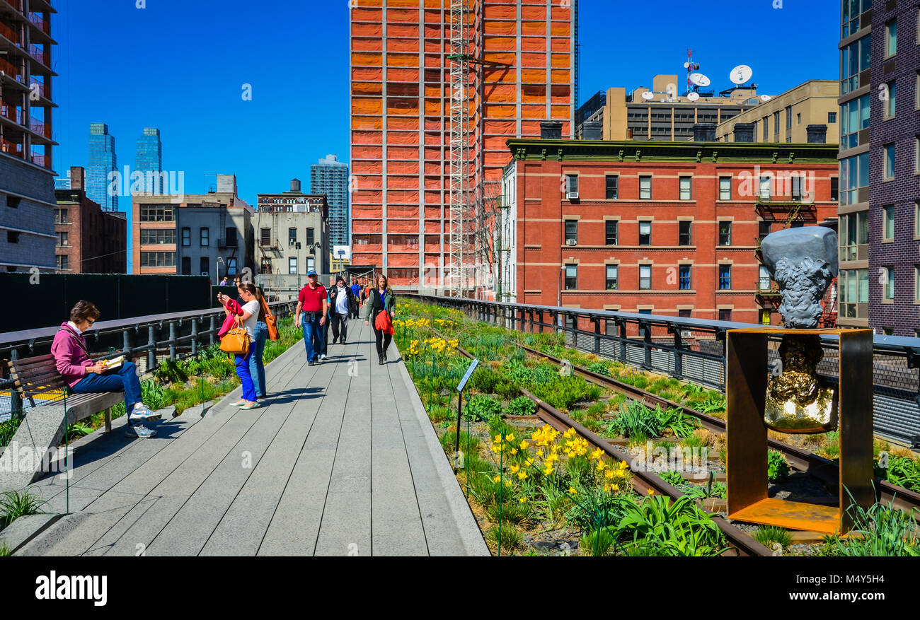 A golden sculpture, a woman sitting on a bench and reading a book, tourists taking photos, and pedestrians walking along path in High Line Park, NYC. Stock Photo