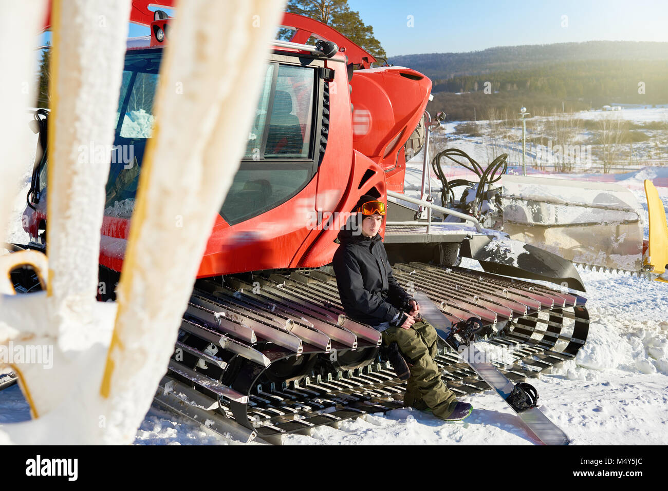 Full length portrait of modern young snowboarder sitting on caterpillar drives of snowcat at snow covered piste of ski resort, copy space Stock Photo