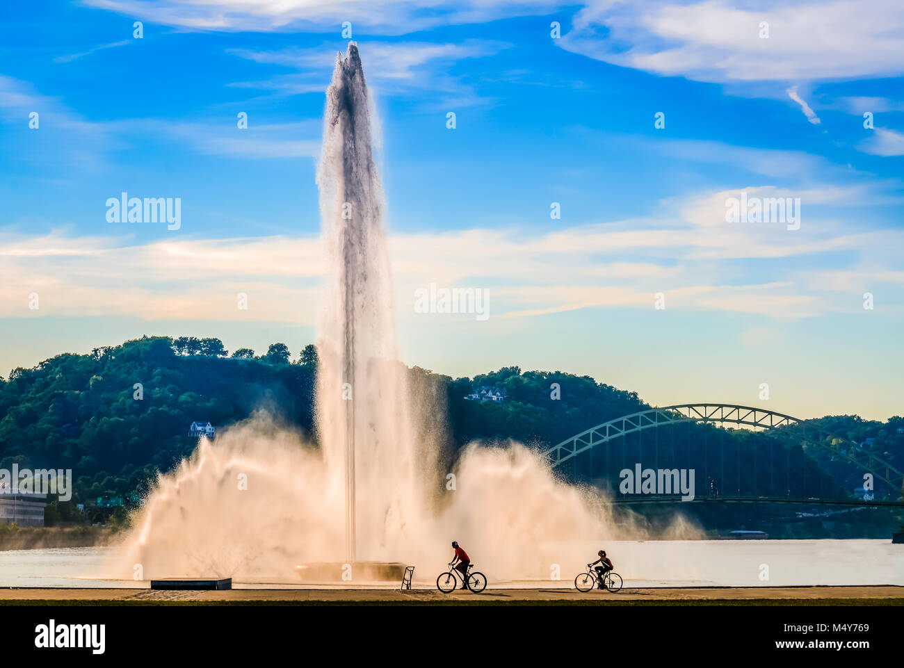 Two bicyclists pass water fountain at Point State Park along Ohio River in Pittsburgh, PA. Stock Photo