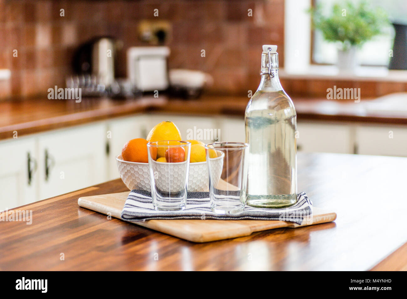 Kitchen interior with table decoration - glass water bottle with stopper, bowl of fruits and glasses, wooden chopping board, tea towel. Home staging. Stock Photo