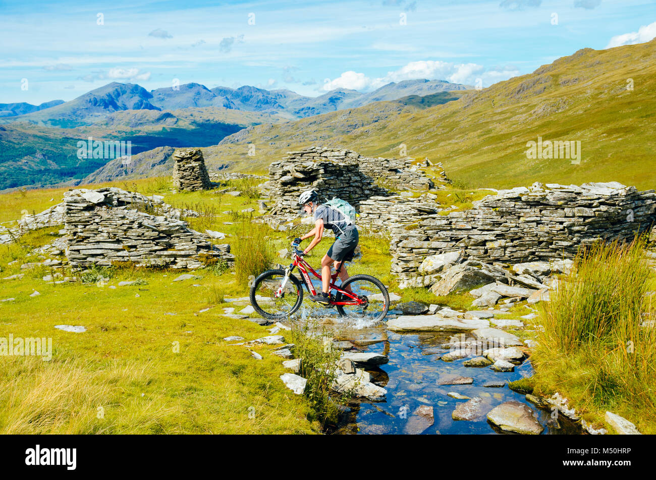 Female mountain biker passing quarry ruins at Walna Scar Quarries in the English Lake District with Scafell and Scafell Pike in the distance Stock Photo