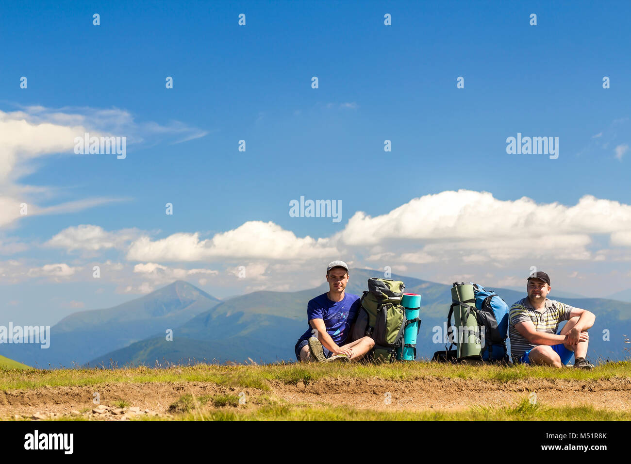Two hikers with backpacks sitting on a hill with mountains in view ...