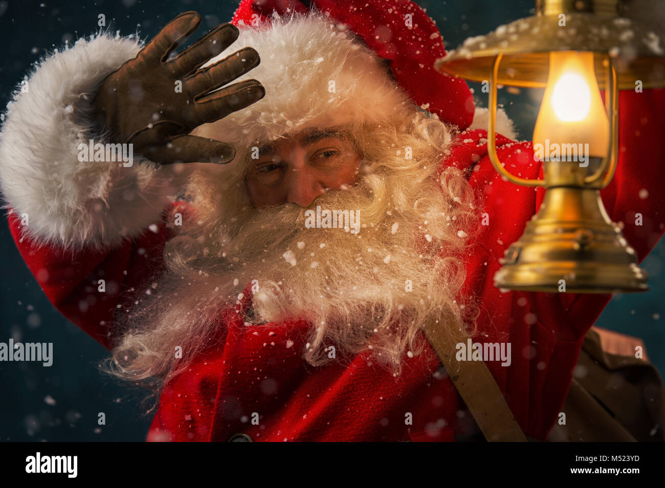 Portrait of happy Santa Claus holding sack with gifts and walking under snowfall with vintage lantern outdoors Stock Photo