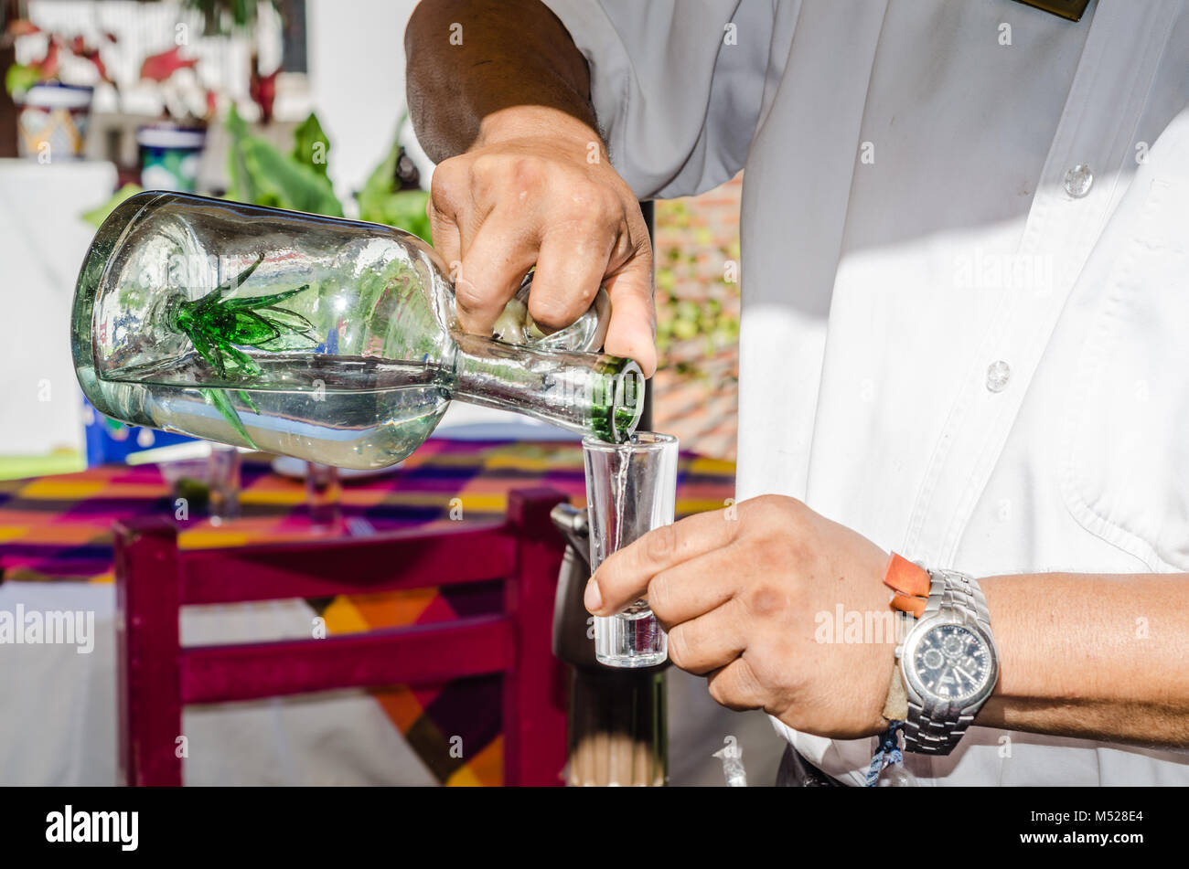 Waiter pours tequila shot from a transparent glass bottle with an agave plant inset in the bottle. Stock Photo