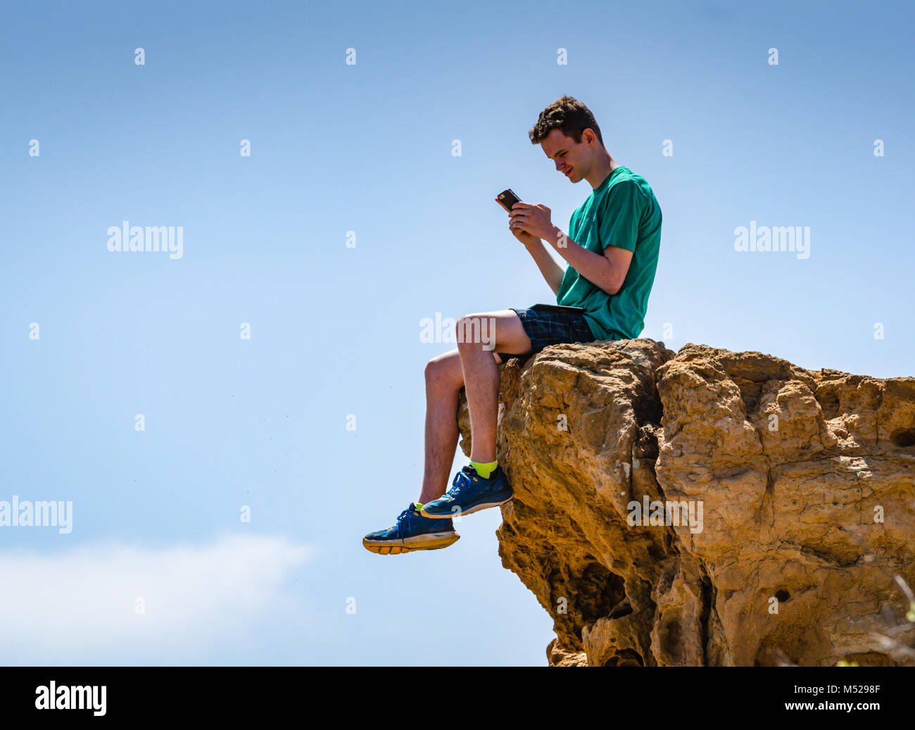 Teen boy sitting on a cliff and reading smartphone in Torrey Pines State Natural Preserve near San Diego, CA. Stock Photo