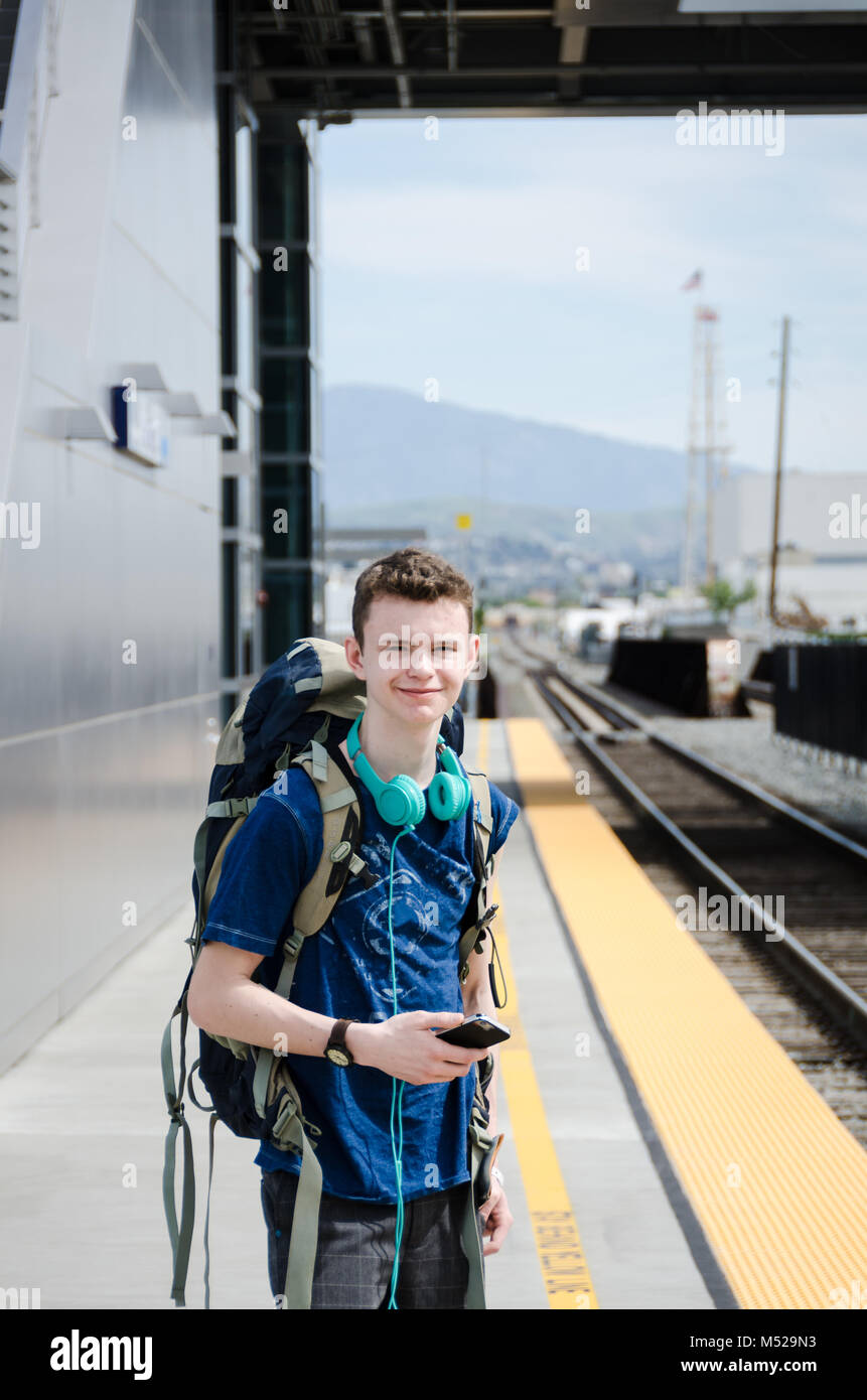 Teenaged boy,  with a backpack on his back, headphones around his neck and holding a smartphone, awaits the train at Anaheim Station. Stock Photo