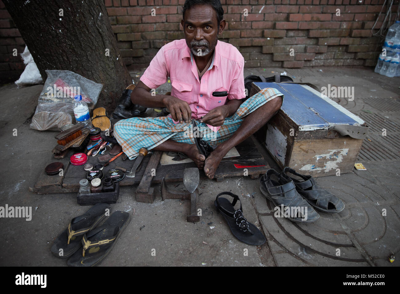 A cobbler sits working with his hands and his feet at the side of the street.  Dhaka, the capital of Bangladesh lies along the east bank of the Buriganga River in the heart of the Bengal delta. The city is a microcosm of the entire country, with diverse religious and ethnic communities. Dhaka is the economic, cultural and political center of Bangladesh and is a major financial center of South Asia. It is one of the world's most populated cities with a population of 18.89 million people in the Greater Dhaka Area. Stock Photo