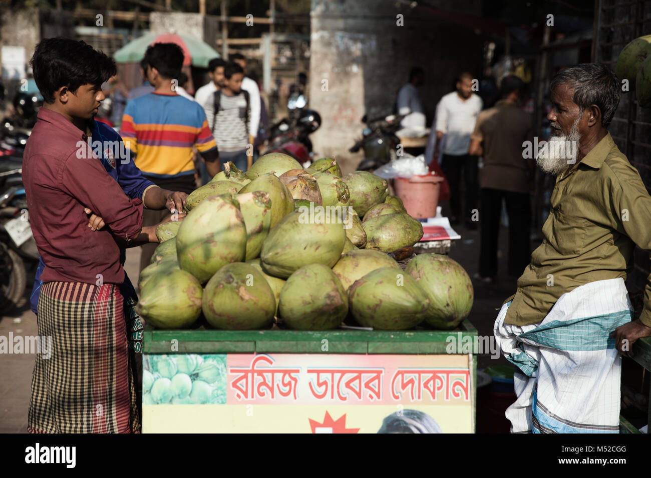 Two men talk business over a coconut seller's stall.  Dhaka, the capital of Bangladesh lies along the east bank of the Buriganga River in the heart of the Bengal delta. The city is a microcosm of the entire country, with diverse religious and ethnic communities. Dhaka is the economic, cultural and political center of Bangladesh and is a major financial center of South Asia. It is one of the world's most populated cities with a population of 18.89 million people in the Greater Dhaka Area. Stock Photo