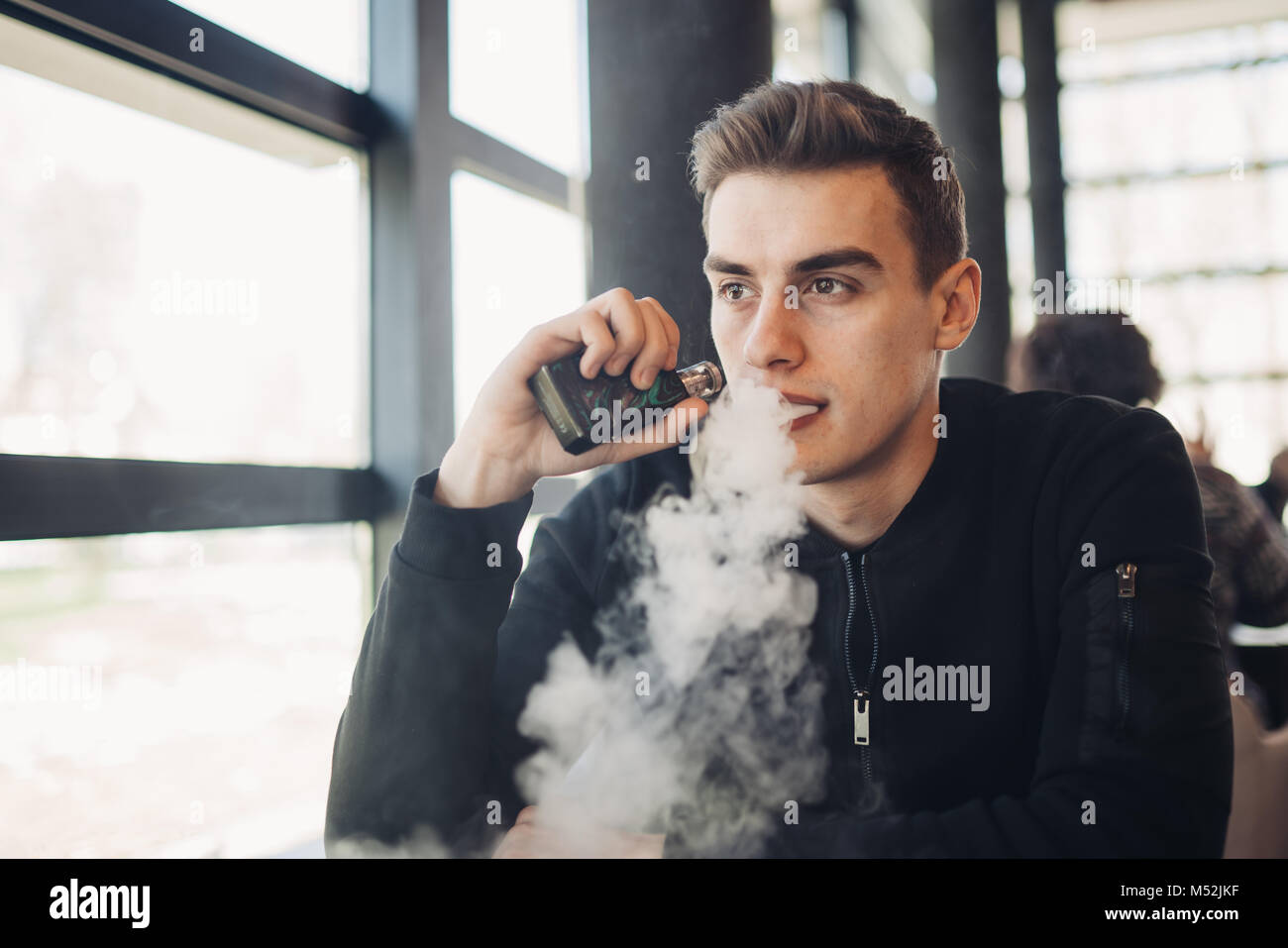 Young man vaping in closed public space.Smoking electronic cigarette in cafe.Nicotine addiction.Way to quit smoking,old habit.Vaping aroma,urban man u Stock Photo