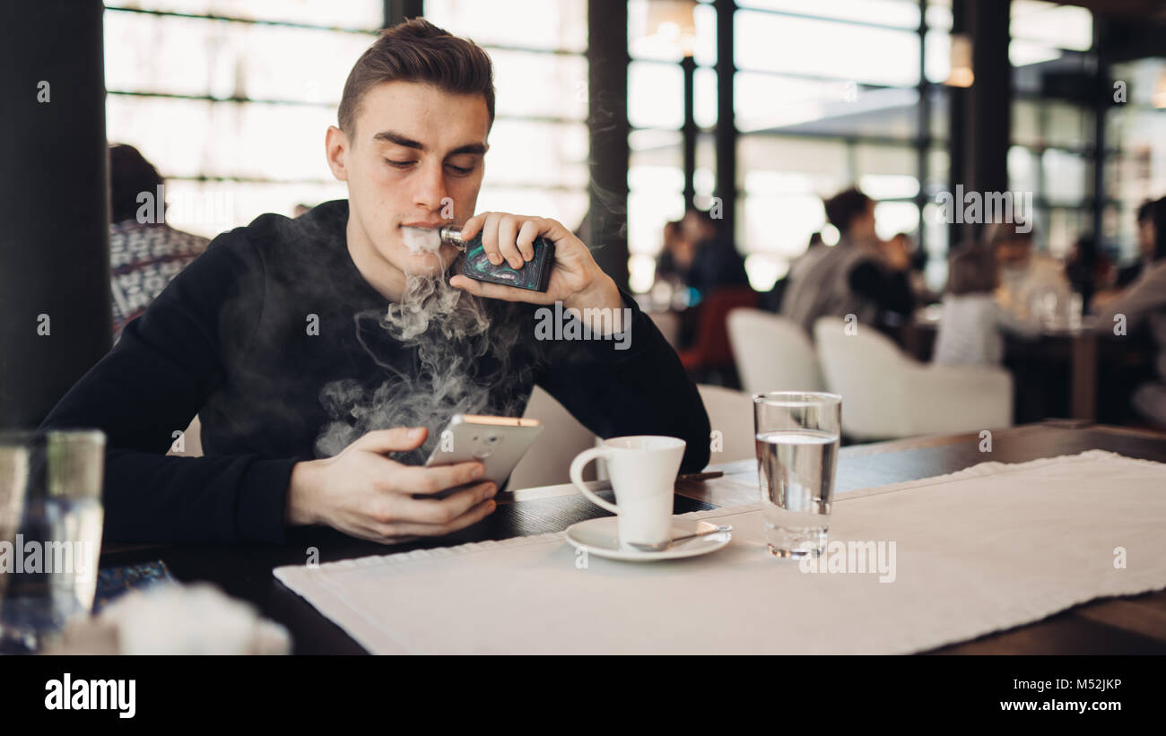 Young man using electronic cigarette to smoke in closed public space.Satisfied e- cigarette user in cafe.Smoking ban alternative,nicotine addiction,qu Stock Photo
