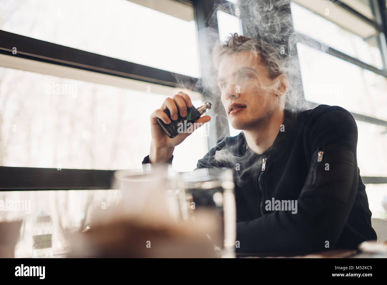 Young man vaping in closed public space.Smoking electronic cigarette in cafe.Nicotine addiction.Way to quit smoking,old habit.Vaping aroma,urban man u Stock Photo