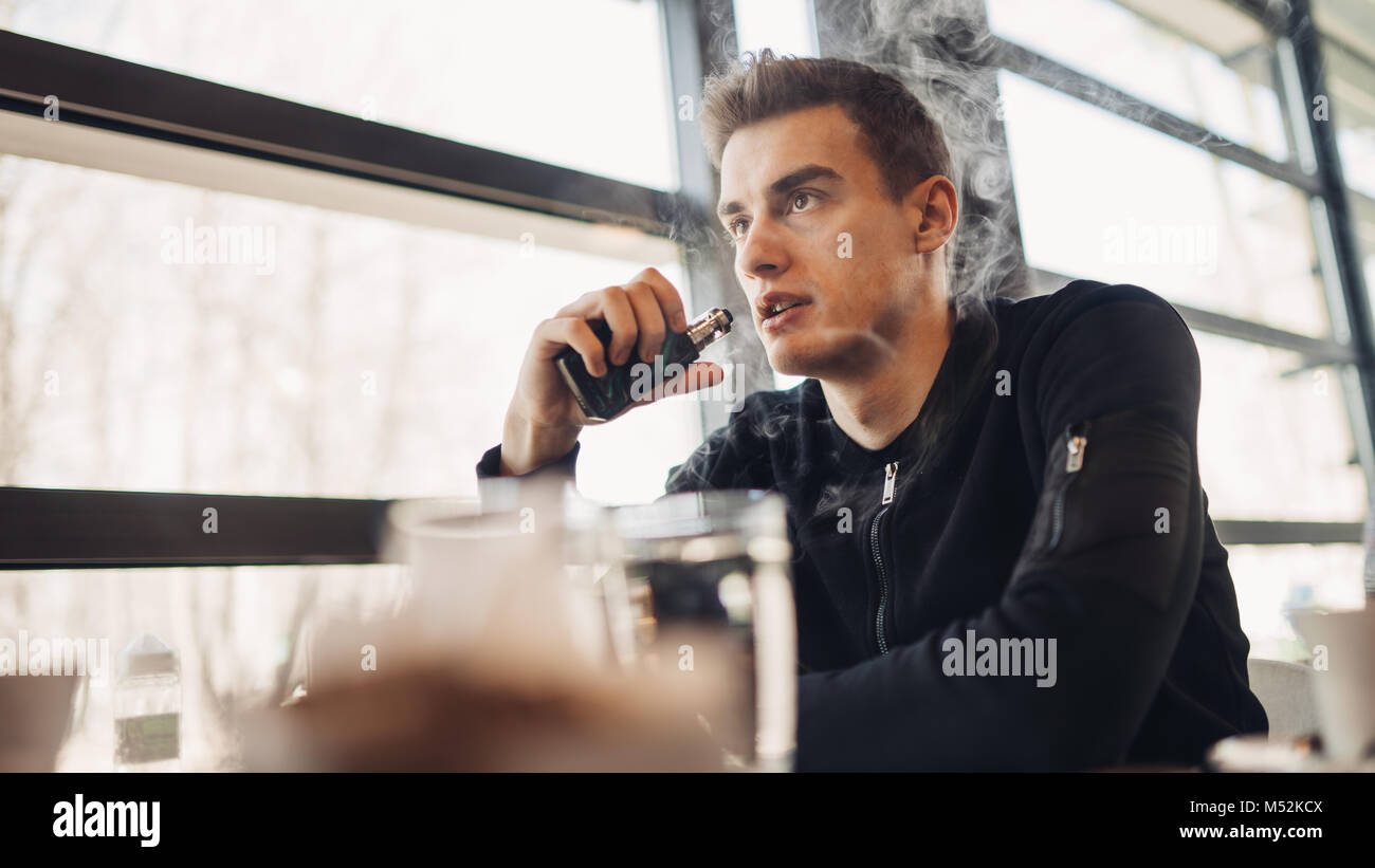 Young man vaping in closed public space.Smoking electronic cigarette in cafe.Nicotine addiction.Way to quit smoking,old habit.Vaping aroma,urban man u Stock Photo