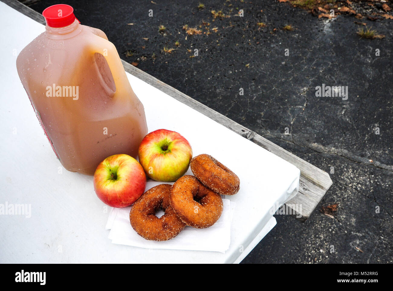 Cold pressed apple cider in a plastic gallon jug dotted by moisture drops of condensation next to two Gala apples and three cider donuts. Stock Photo
