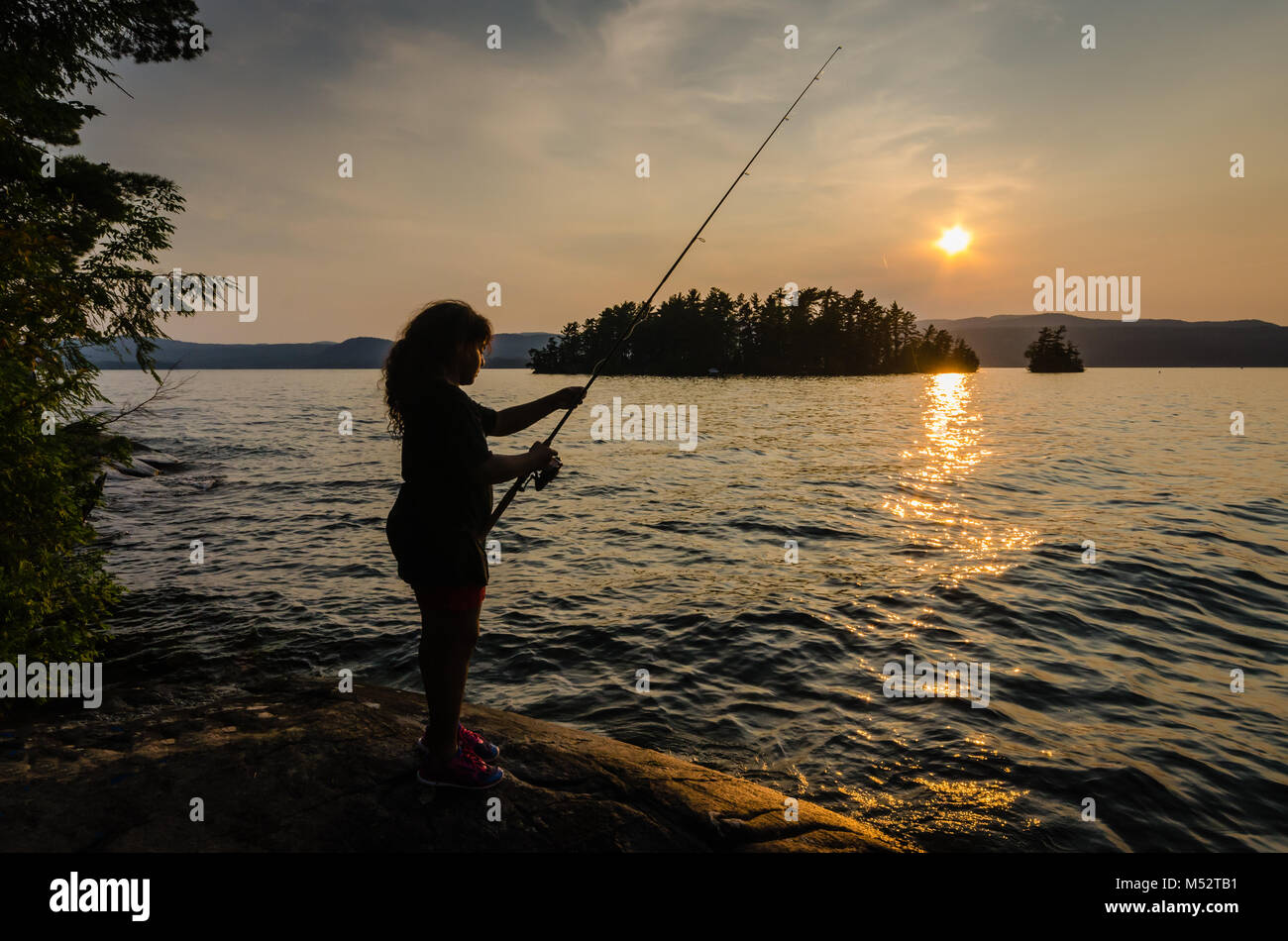 Young Hispanic girl fishes on a rock at Lake George in New York near the Adirondack Mountains as the setting sun casts a golden glow over her face. Stock Photo