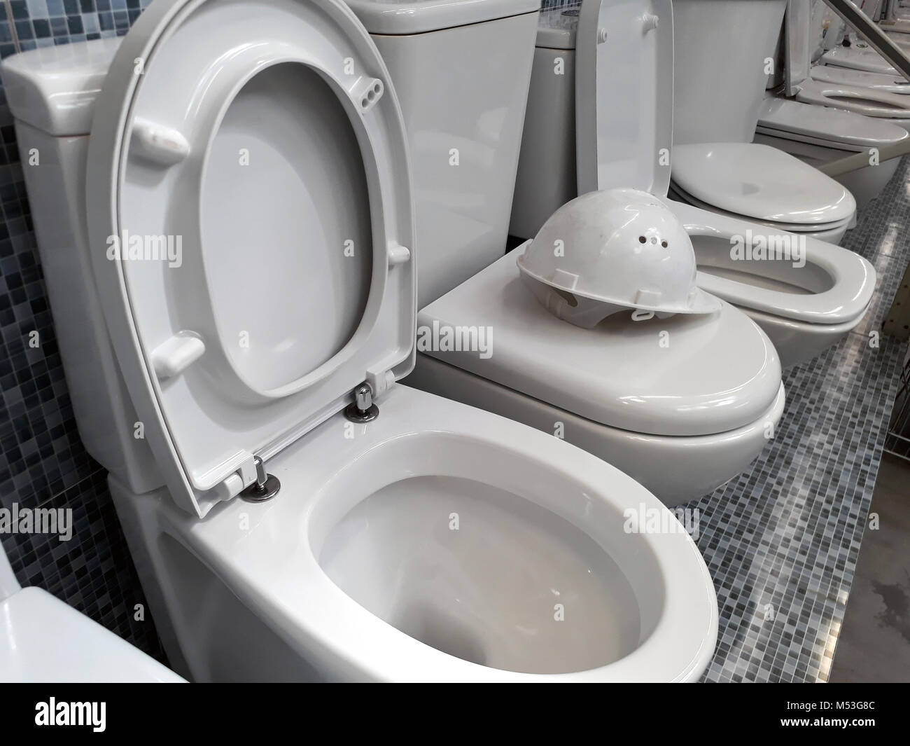 A number of white toilet bowls in the store and white construction helmet on top of one of them Stock Photo
