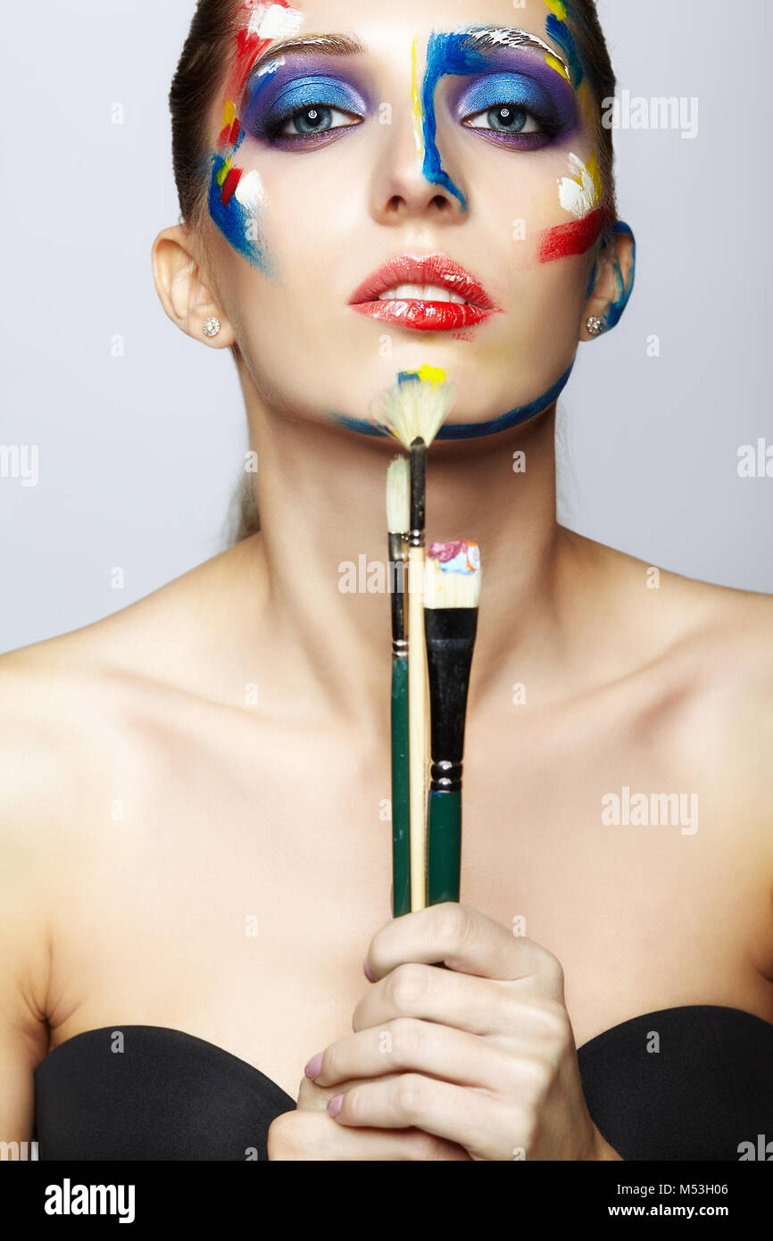 Young woman painter with paint brush in hand and acrylic paint on face ...